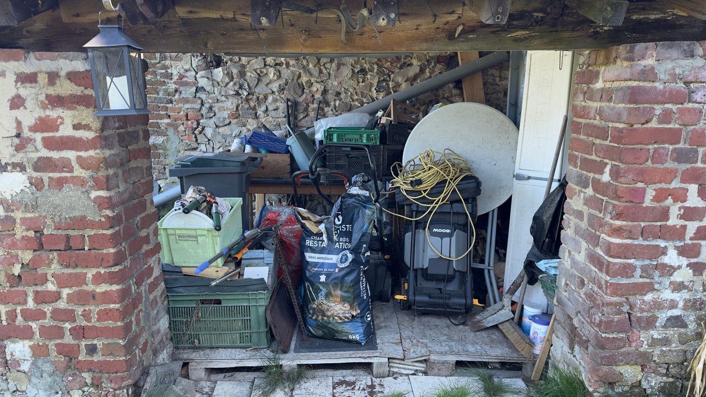 Appentis encombré de bacs, d'outils de jardin, de sacs et de mobilier avant l'intervention